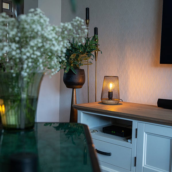 Dekorative Gittertischleuchte mit Holzfuß auf einem Sideboard in einem gemütlich eingerichteten Wohnzimmer mit Zimmerpflanze.