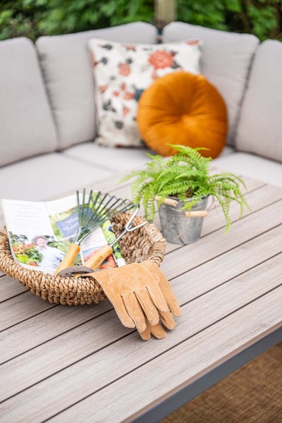 Holztisch mit geflochtenem Korb, Gartenwerkzeug, Lederhandschuhen und Pflanze vor einer grauen Lounge mit Kissen.