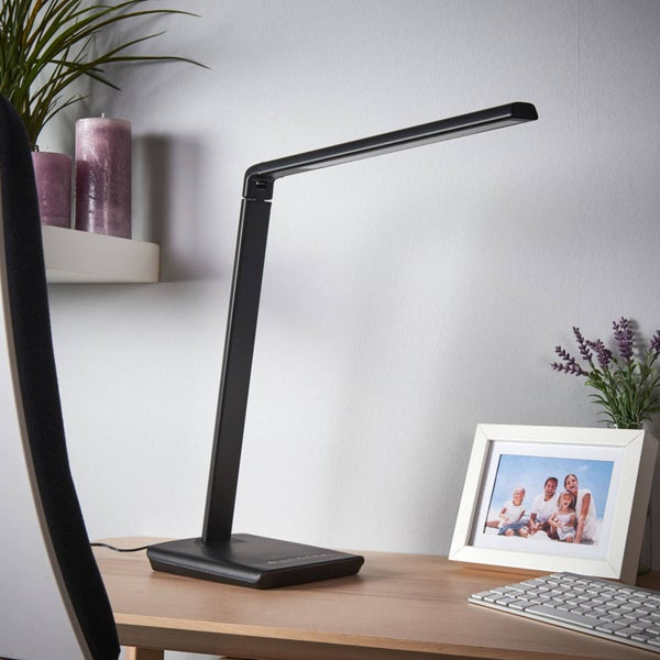 Moderne schwarze LED Schreibtischleuchte mit verstellbarem Arm auf einem Holzschreibtisch neben Tastatur, Bilderrahmen und kleiner Pflanze.