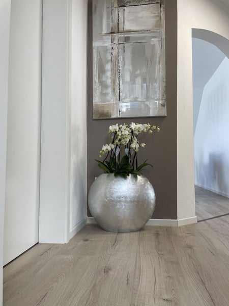 Modernes Interieur mit rundem silbernen Pflanzgefäß, weißen Orchideen und Holzboden unter einem abstrakten Gemälde.