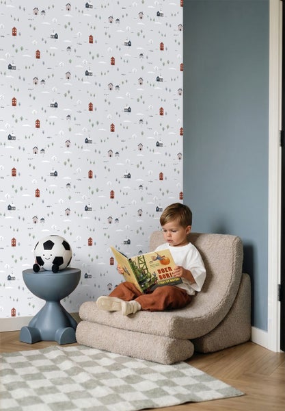 Helles Kinderzimmer mit einer Tapete mit Hausmotiven, einem Polstersessel, einem Beistelltisch mit Spielzeug und einem lesenden Kind.