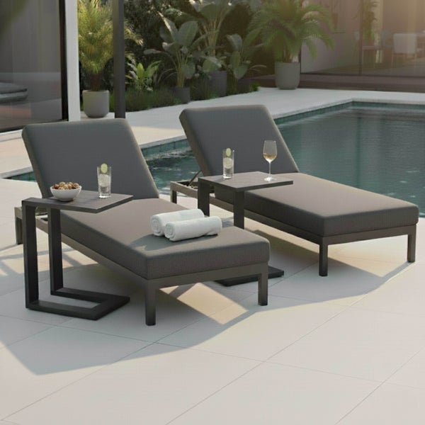 Zwei moderne graue Sonnenliegen mit passenden Beistelltischen auf einer Terrasse am Pool mit tropischen Pflanzen.
