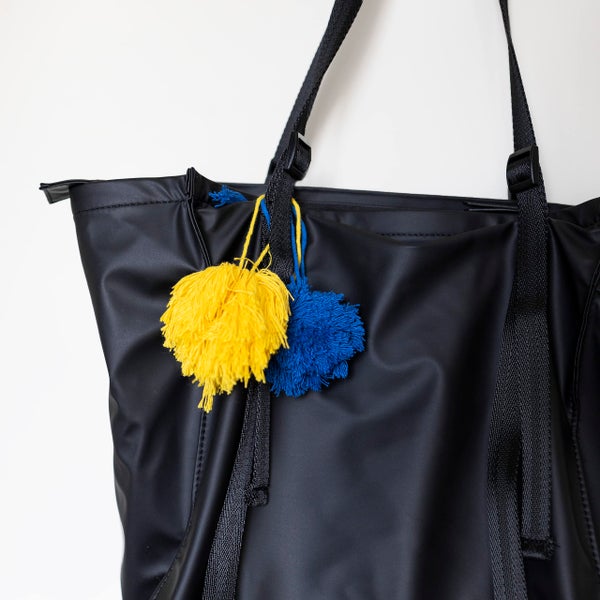 Schwarze Tragetasche aus Kunststoff mit verstellbaren Trageriemen und gelben sowie blauen Bommel-Anhängern.