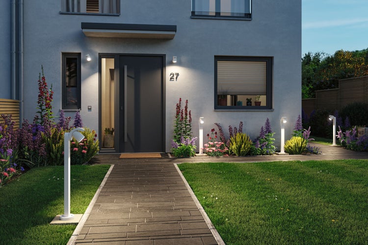 Moderner Hauseingang bei Dämmerung mit beleuchtetem Gehweg, Außenleuchten in den Beeten und an der Fassade, gepflegter Rasen und Blumenbeete.