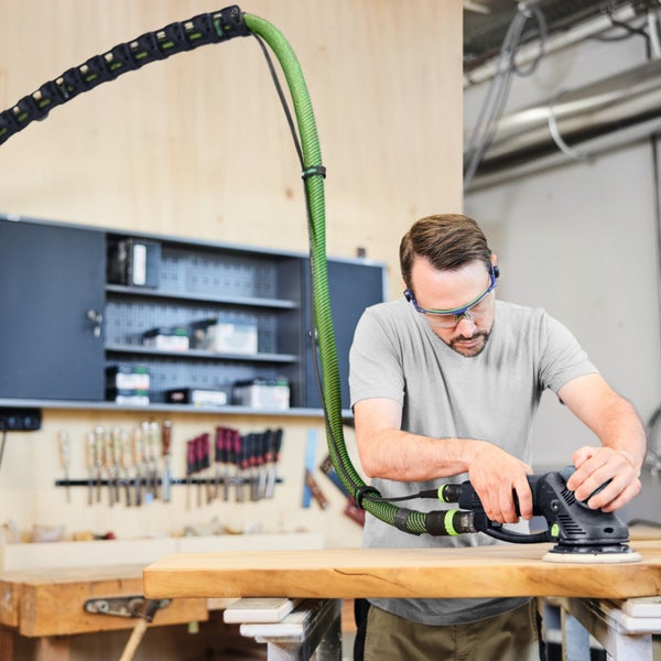 Mann mit Schutzbrille schleift Holzplatte in einer Werkstatt mit einem Exzenterschleifer und angeschlossenem Absaugschlauch.