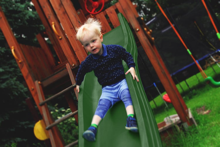 Kleinkind rutscht auf einer grünen Kunststoffrutsche an einem hölzernen Spielturm im Garten.