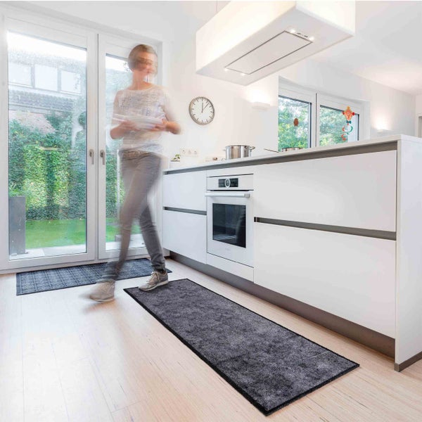 Moderne Küche mit weißen Küchenschränken und zwei grauen Küchenläufern auf dem Holzboden sowie einer Person in Bewegung.