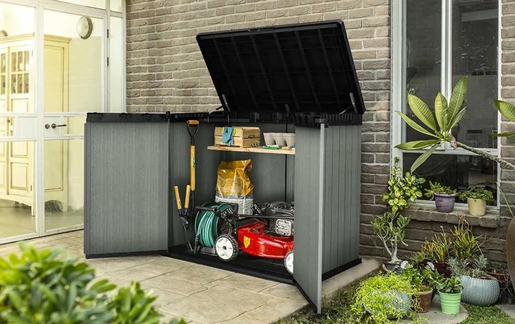 Offener Gartenschrank aus Kunststoff mit Rasenmäher, Gartenschlauch und Werkzeugen vor einer Ziegelwand auf einer Terrasse.