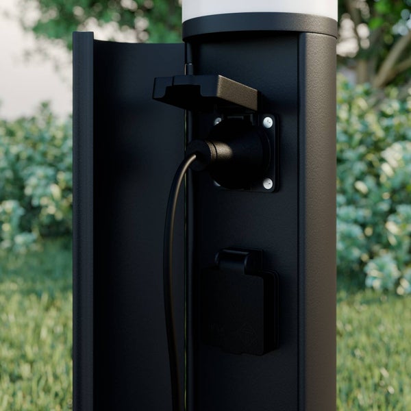 Schwarze Außenleuchte mit zwei integrierten Steckdosen hinter einer Schutzklappe, eine Steckdose mit eingestecktem Kabel.