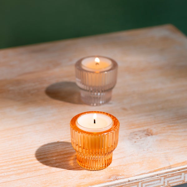 Zwei geriffelte Teelichthalter aus Glas mit brennenden Kerzen auf einem Holztisch.