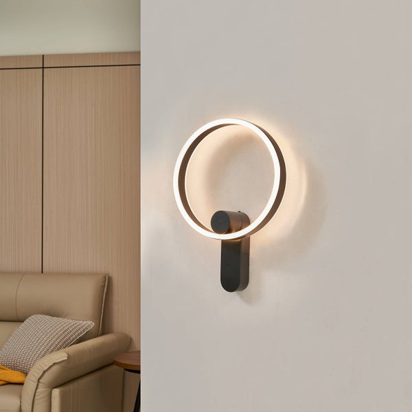 Moderne LED-Wandleuchte in Ringform, schwarz, montiert an einer hellen Wand in einem Wohnraum mit Sofa und Holzpaneelen.