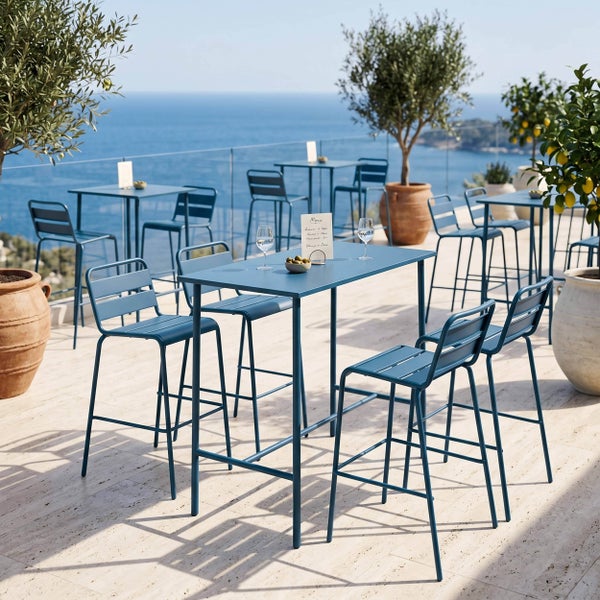 Blaue Stehtische und Barstühle aus Metall auf einer hellen Terrasse mit Meerblick und Olivenbäumen in Terrakottakübeln.