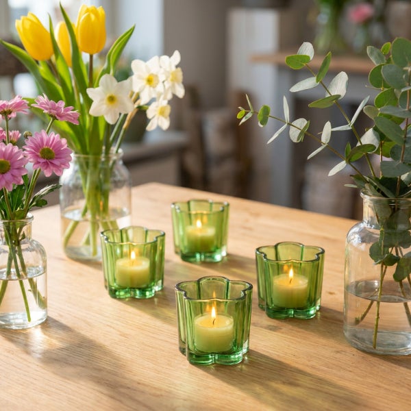 Tischdekoration mit drei blumenförmigen Glas Teelichthaltern und zwei Glasvasen mit Frühlingsblumen und Eukalyptus auf einem Holztisch.