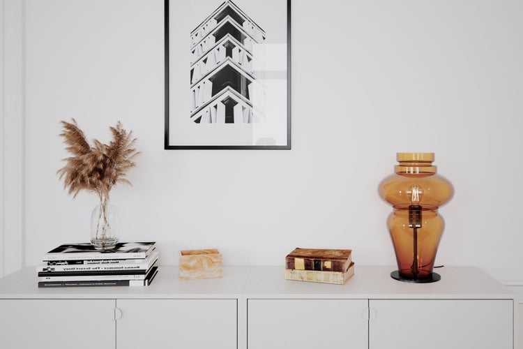 Dekoratives Wohnambiente mit Tischleuchte, Trockenblumen, Bücherstapel und einem gerahmten Bild an der Wand über einem weißen Schrank.