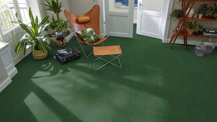 Grüner Teppichfliesenboden in einem hellen Wohnzimmer mit Schmetterlingsstuhl aus Leder, Zimmerpflanzen und Holzregal.