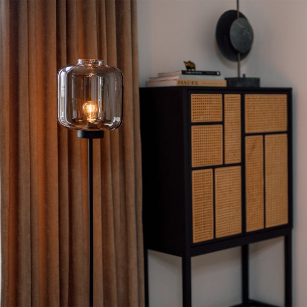 Stehlampe mit transparentem Lampenschirm und schwarzem Gestell neben einem Schrank mit Rattangeflecht