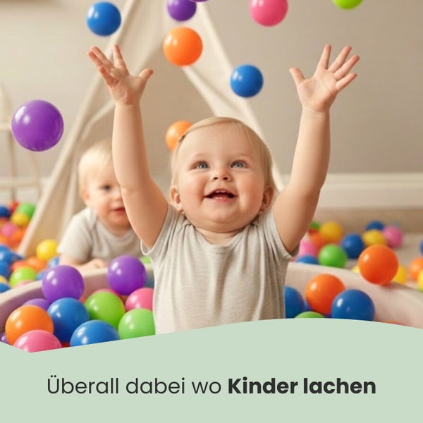Ein lachendes Baby sitzt in einem Bällebad mit bunten Spielbällen und hebt die Arme. Im Hintergrund steht ein helles Tipi-Zelt.
