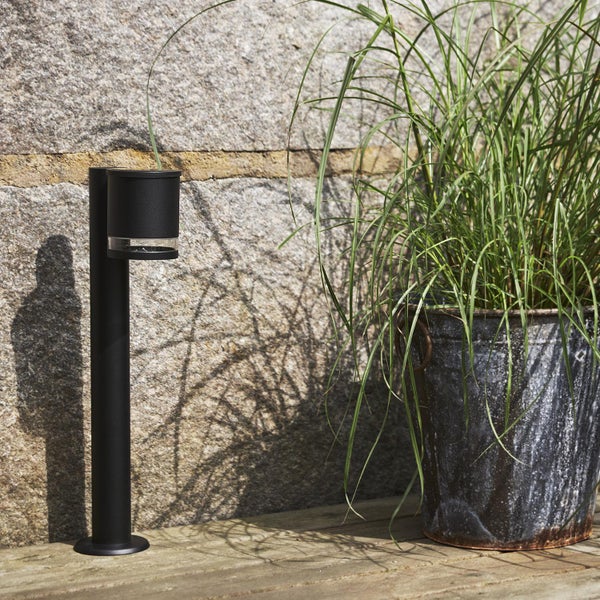 Eine schwarze Wegeleuchte neben einer Pflanze im Topf vor einer Steinwand.