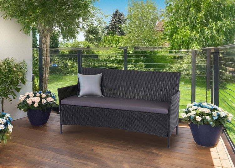 Dunkelgraue Polyrattan Gartenbank mit Sitzkissen und Zierkissen auf einer Holzterrasse mit Blumenkübeln und Garten im Hintergrund.