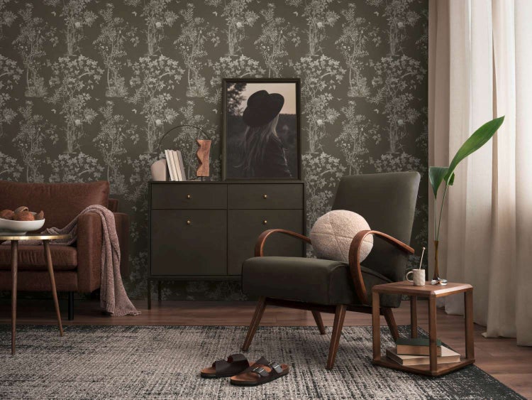 Wohnzimmer mit Vliestapete mit botanischem Muster, beigem Sessel mit Holzarmlehnen, Kommode, Teppich und Einrichtungsgegenständen.