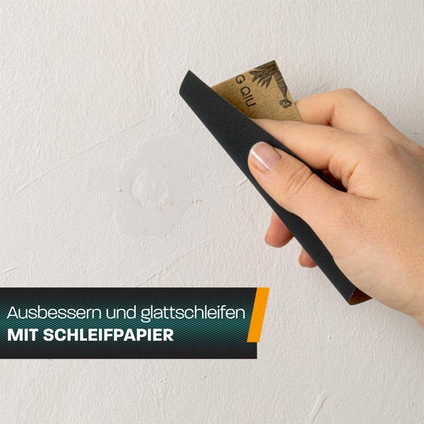 Hand beim Glattschleifen einer ausgebesserten Stelle an einer Wand mit einem Blatt Schleifpapier.