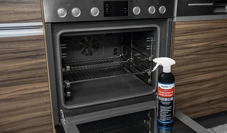 Sprühflasche mit Backofenreiniger und Grillreiniger steht vor einem geöffneten Einbaubackofen in einer Küche.
