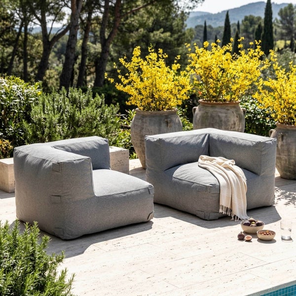 Zwei graue Outdoor-Loungesessel im Sitzsack-Stil auf einer hellen Steinterrasse, umgeben von großen Pflanzgefäßen mit gelben Blumen und Bäumen.