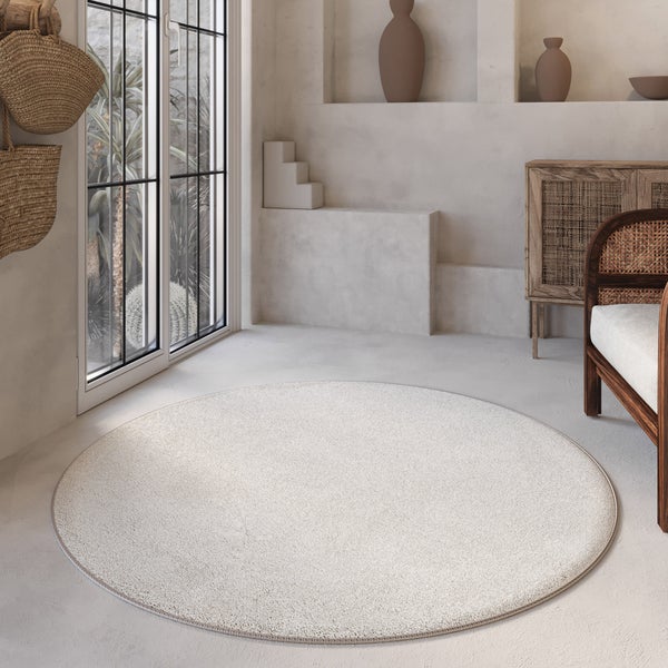 Runder cremefarbener Teppich in hellem, minimalistischem Wohnraum mit Holzmoebeln, Geflecht-Elementen und grossen Fenstern.