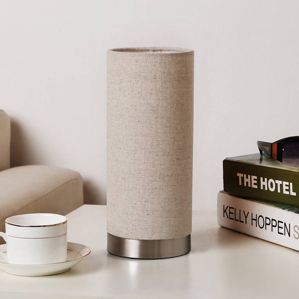 Zylindrische Tischleuchte mit beigem Textilschirm und gebürstetem Metallsockel auf einem Tisch neben einer Tasse und Büchern.