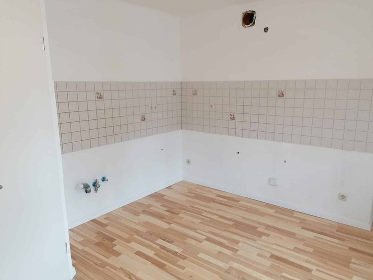Renovierter leerer Raum mit Laminatboden in heller Holzoptik, weißen Wänden und einem Fliesenspiegel für eine Küche.
