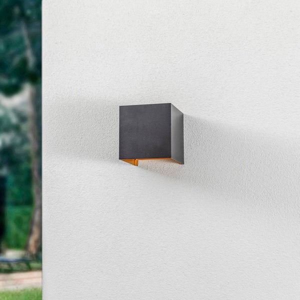 Würfelförmige Außenwandleuchte in Anthrazit an einer weißen Hauswand mit warmem Lichtaustritt nach unten.