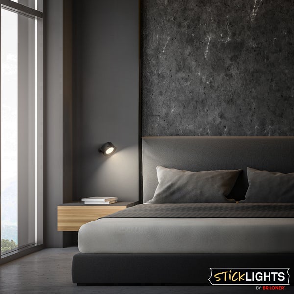 Modernes Schlafzimmer mit schwarzer Wandleuchte, dunkler Strukturwand und grauem Polsterbett. Sticklights by Briloner Logo.