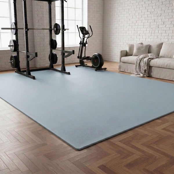 Bodenschutzmatte für Fitnessgeräte in einem hellen Raum mit Hantelstation, Crosstrainer und Sofa auf Parkettboden im Fischgrätenmuster.