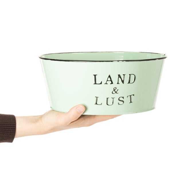 Eine Hand hält eine hellgrüne Pflanzschale aus Metall mit der Aufschrift Land und Lust.