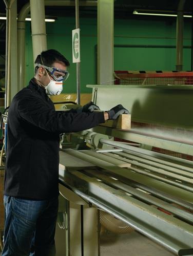 Ein Arbeiter mit Schutzbrille und Staubmaske bedient eine Holzbearbeitungsmaschine in einer Werkstatt.