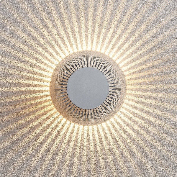 Moderne LED-Wandleuchte mit rundem Gehäuse, die ein radiales Strahlenmuster aus Licht an eine strukturierte Wand wirft.
