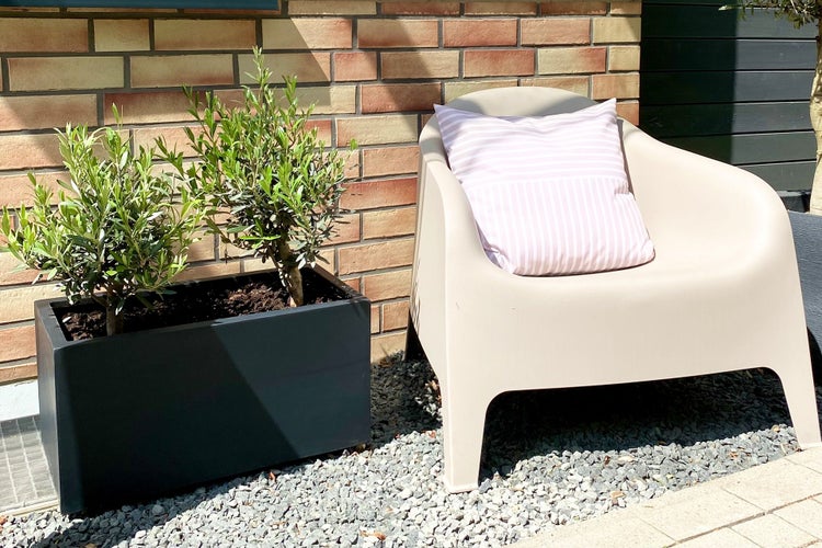 Beiger Gartenstuhl aus Kunststoff mit gestreiftem Kissen neben einem anthrazitfarbenen Pflanzkasten auf Kies vor einer Ziegelwand.