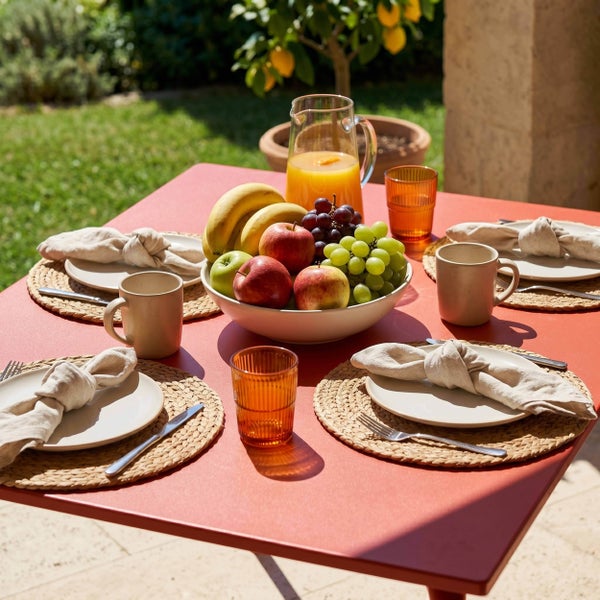 Gedeckter Gartentisch mit Obstschale, Tellern auf geflochtenen Platzsets und Saftkaraffe in einer sonnigen Gartenumgebung.