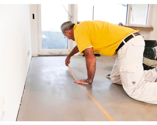 Ein Mann kniet auf dem Boden und klebt Klebeband auf einen Zementestrich.