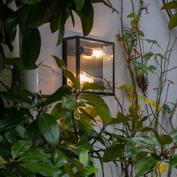 Moderne quadratische Außenwandleuchte mit zwei leuchtenden Filamentlampen an einer Hauswand zwischen grünen Blättern.
