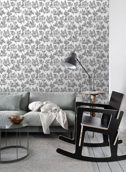 Vliestapete mit Toile-de-Jouy-Muster in einem hellen Wohnzimmer mit grauem Sofa, schwarzem Schaukelstuhl und Tischlampe.