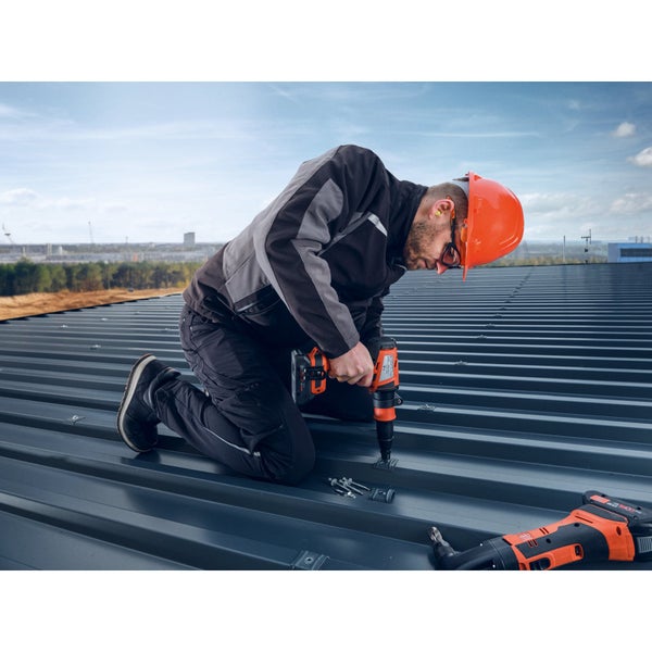 Handwerker mit Schutzhelm und Arbeitskleidung verschraubt Wellblechplatten auf einem Dach mit einem Akku Schrauber.
