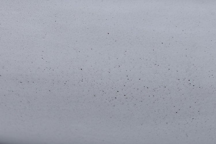 Nahaufnahme einer grauen Betonoberfläche mit feiner poröser Struktur und dunklen Einschlüssen.