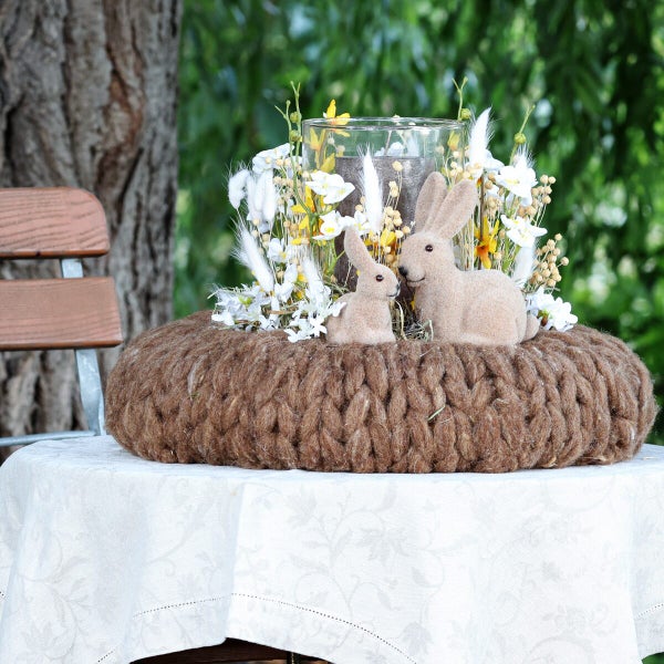 Osterkranz aus Strickoptik mit zwei Hasenfiguren und einer Glaswindlichtschale mit Trockenblumen auf einem Tisch im Garten