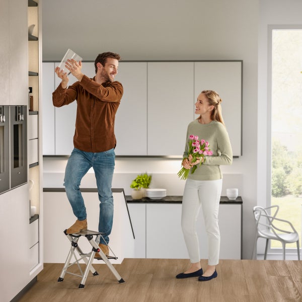 Mann auf Trittstufe in Küche, Frau mit Blumenstrauß