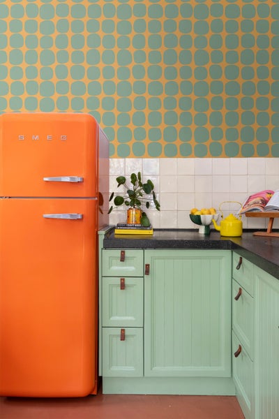 Retro-Küche mit orange gemusterter Tapete, orangefarbenem Kühlschrank und mintgrünen Schränken vor weißem Fliesenspiegel.