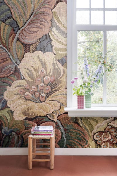 Fototapete mit großflächigem Blumenmuster in Tapisserie-Optik neben einem Fenster mit Blumenvasen und einem Holzhocker.
