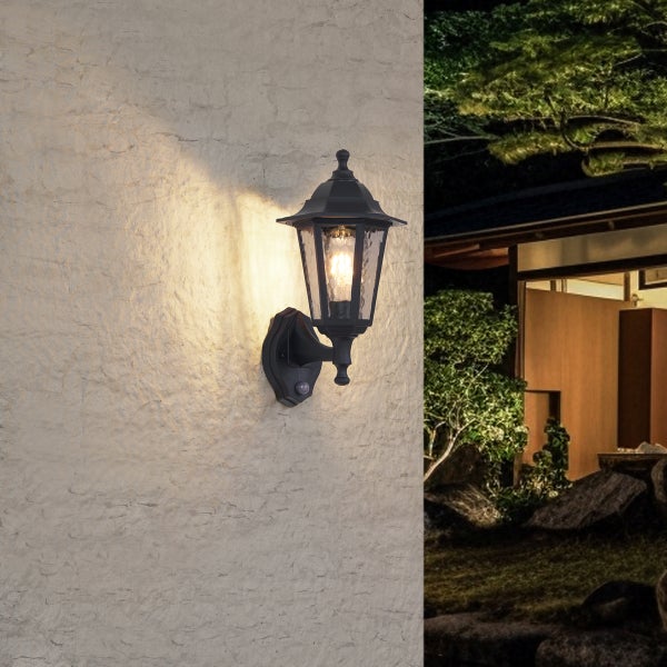 Schwarze Außenwandleuchte im Laternen-Stil mit Bewegungsmelder und strukturiertem Glas an einer hellen Wand, beleuchtet eine nächtliche Szene.