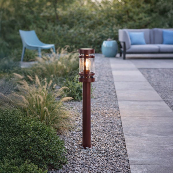 Moderne Wegeleuchte mit warmem Leuchtmittel an einem Gartenweg aus grauen Steinplatten und Kies, umgeben von Gräsern und Sträuchern.