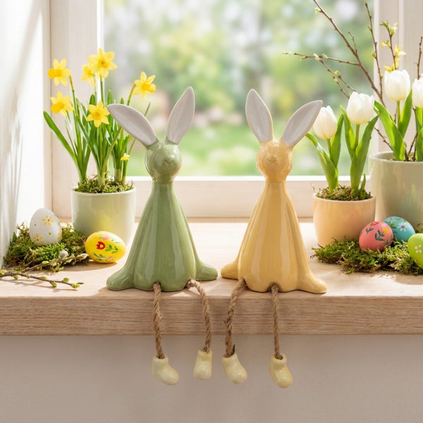 Zwei Osterhasen Figuren aus Keramik mit baumelnden Beinen auf einer Fensterbank dekoriert mit Frühlingsblumen und bemalten Ostereiern.